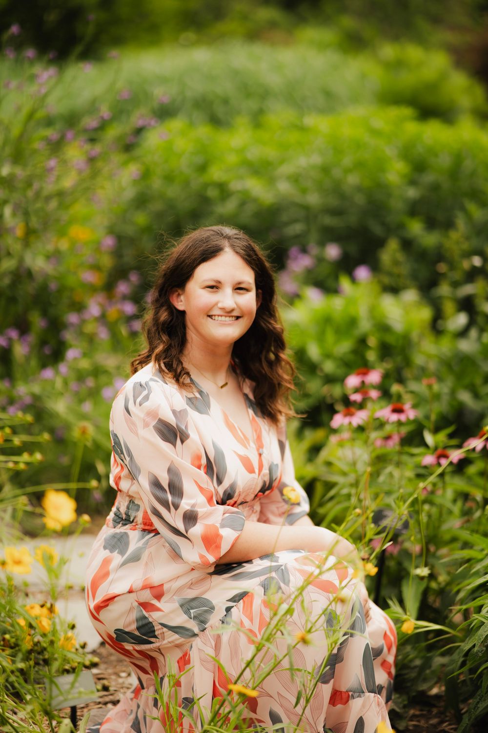 Smiling young woman in peach floral dress sitting among colorful coneflowers in a summer garden.