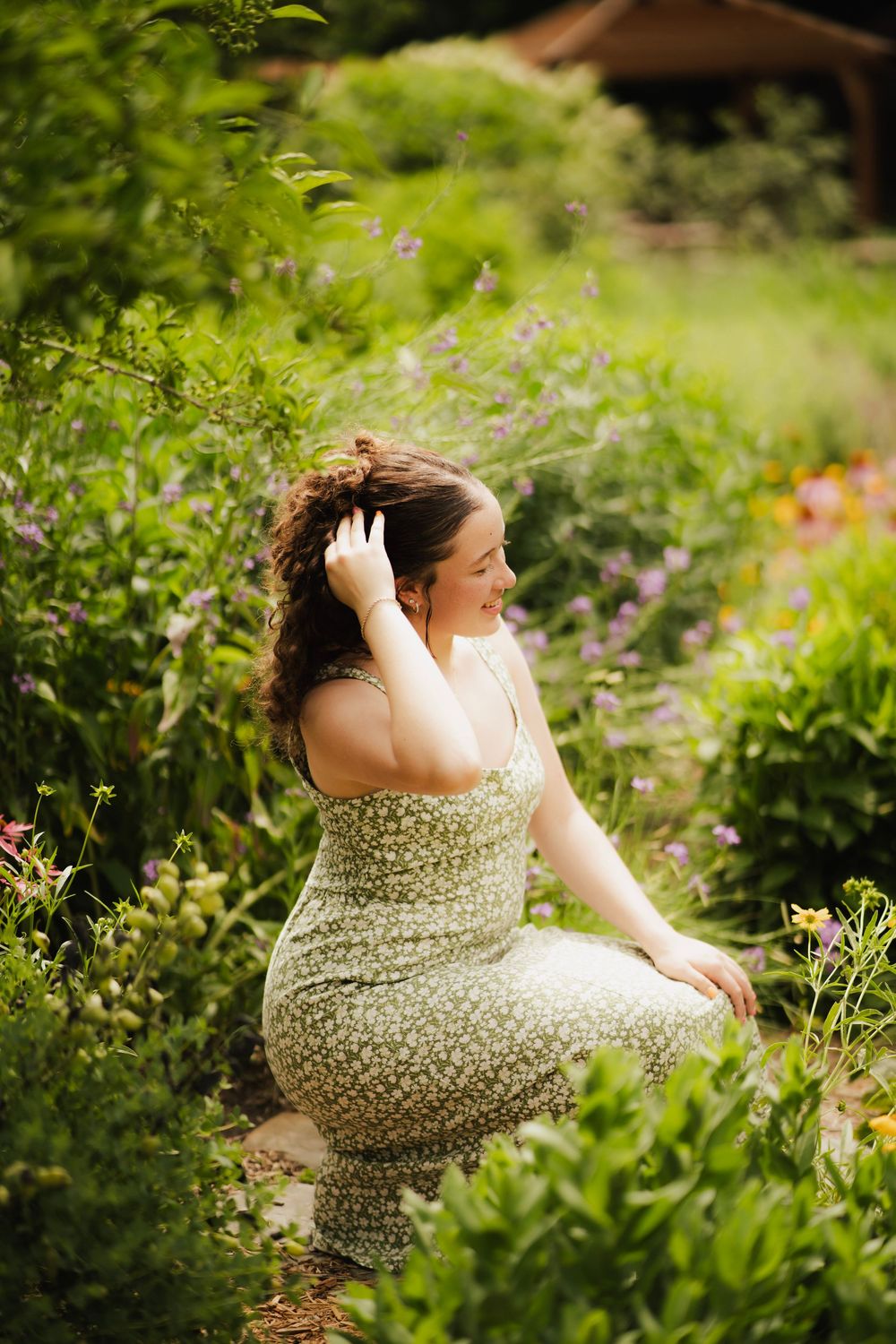 Young woman in green floral sundress kneeling in lush garden, looking to the side smiling.