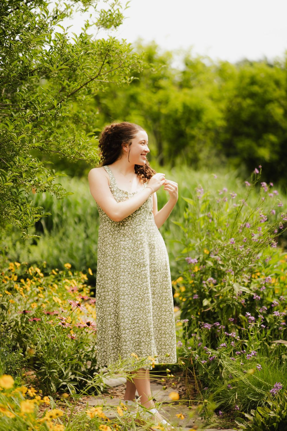 Young woman in green floral sundress smiling and twirling hair in bright sunny garden.