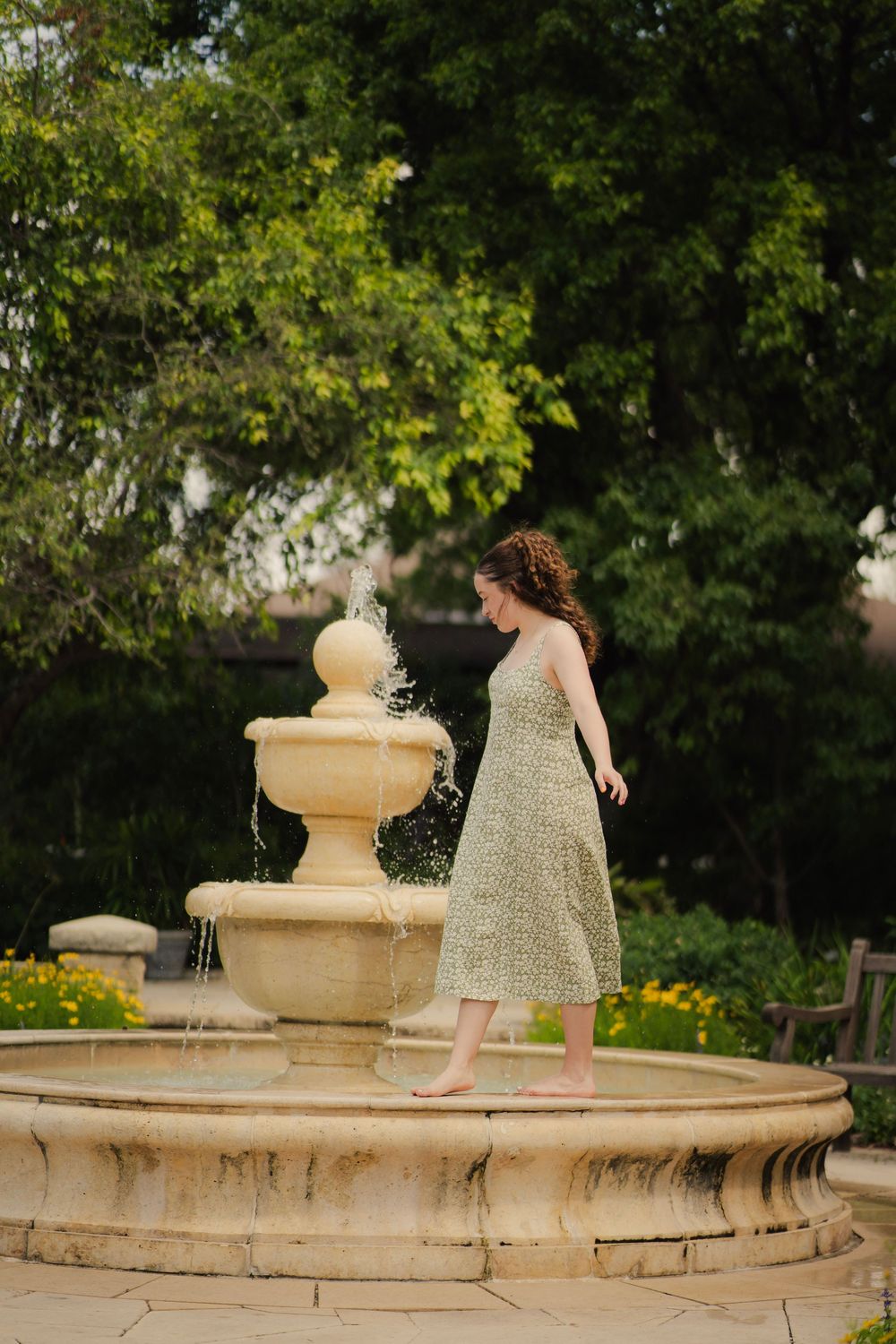 Young woman in green floral midi dress standing barefoot beside a garden fountain.