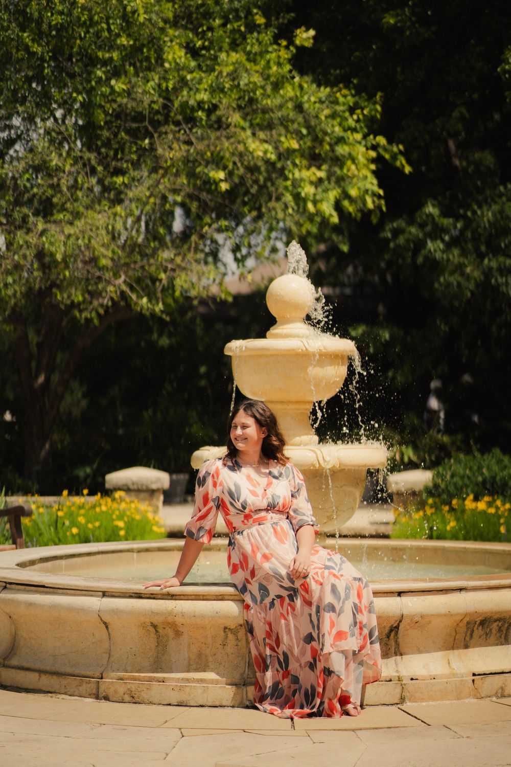 Woman in pink floral maxi dress sitting on stone fountain edge in a sunny garden.