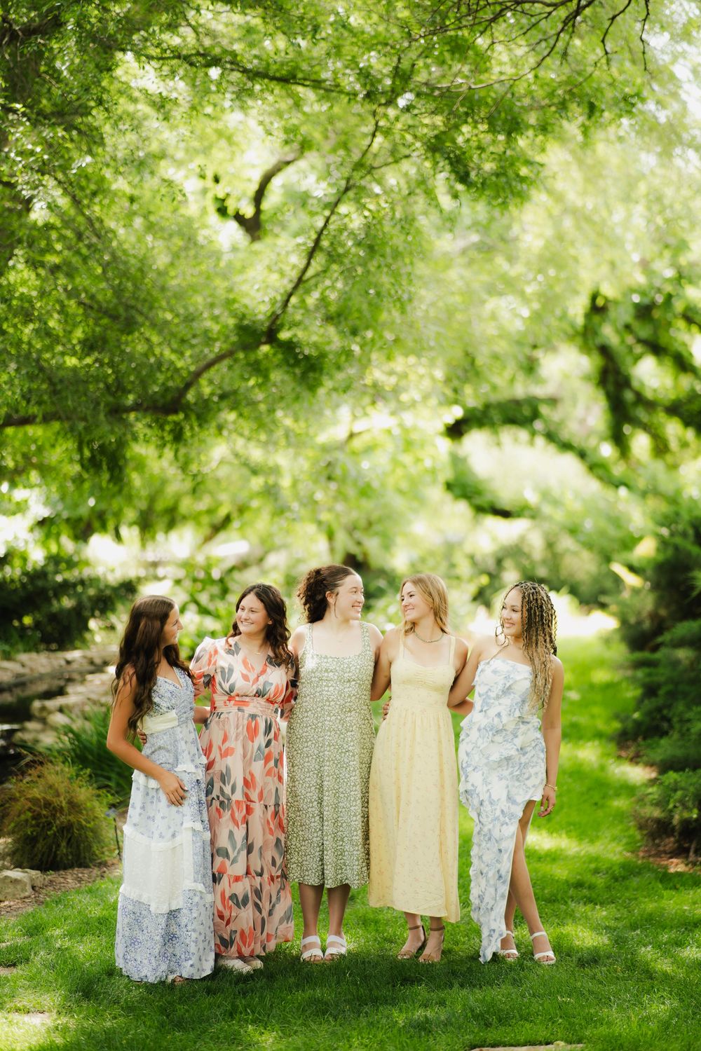 Five women in floral and pastel dresses embracing and smiling under green tree canopy.