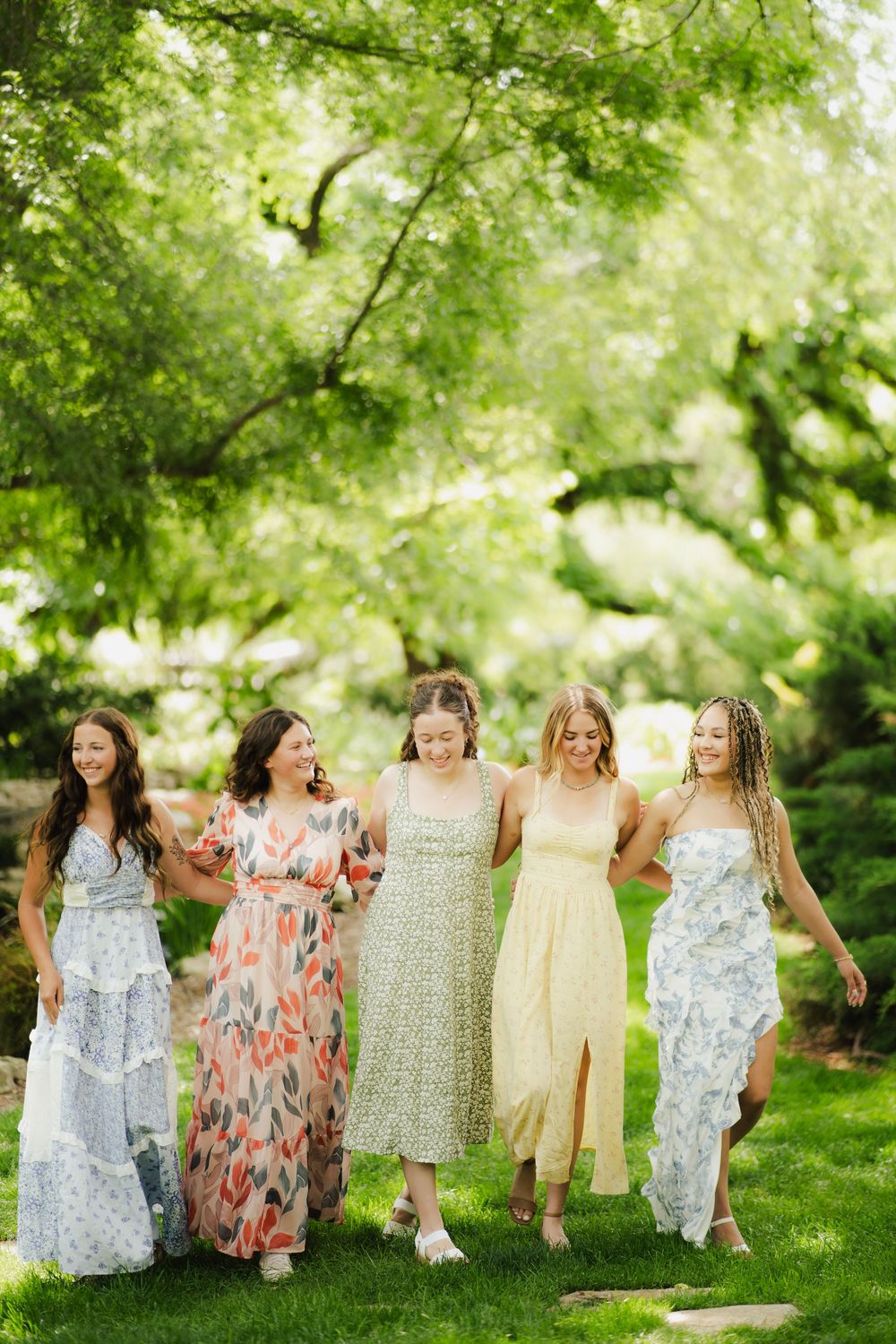Five women in floral and pastel dresses walking arm in arm through a sunny garden.