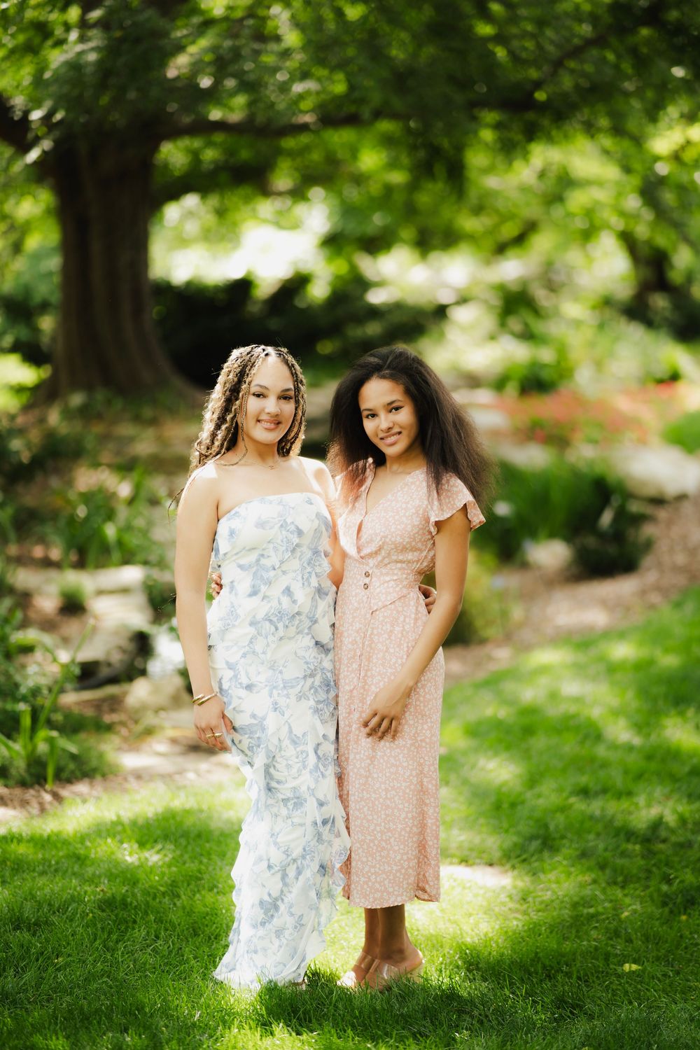 Two young women in floral dresses smiling together in a lush green garden setting.