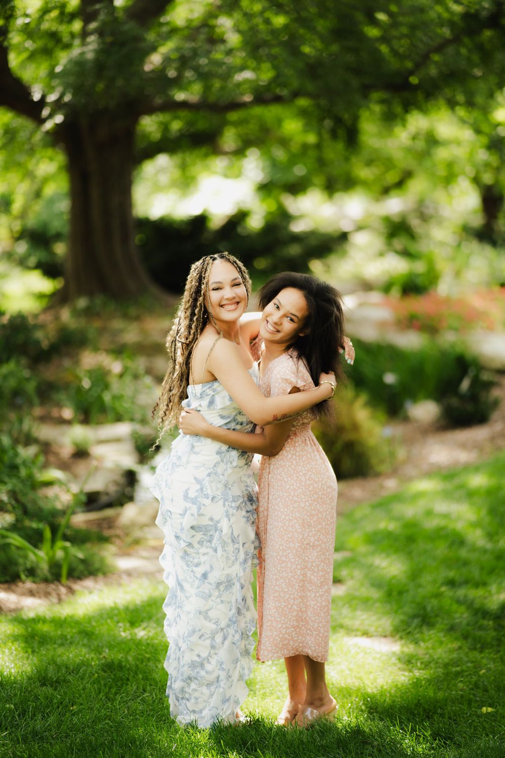 Two young women in floral dresses hugging and smiling in a lush green garden.