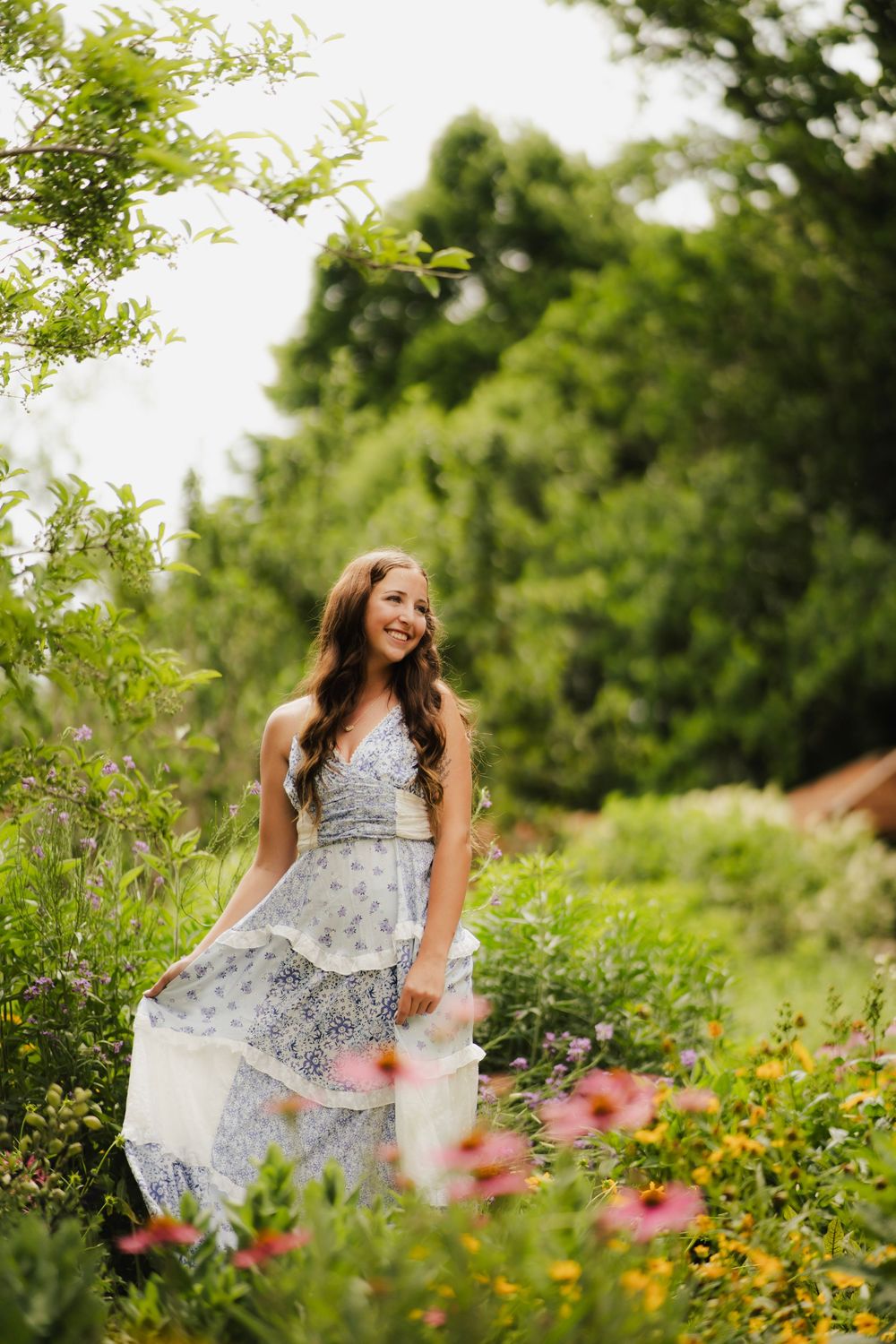 Smiling young woman in blue floral dress laughing among pink and yellow garden flowers.