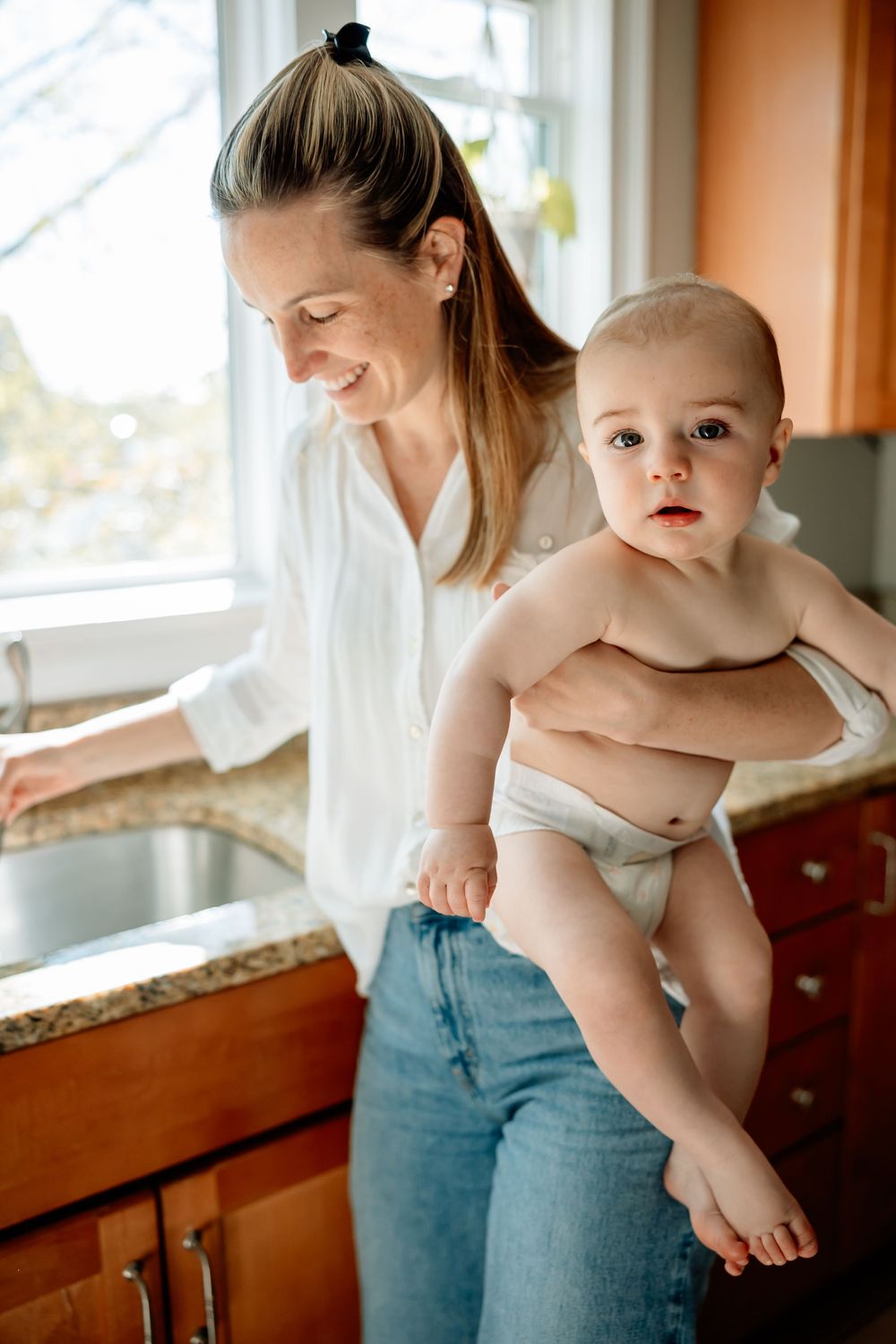 Smiling mother in white shirt and jeans holding baby in diaper near kitchen sink by bright window.