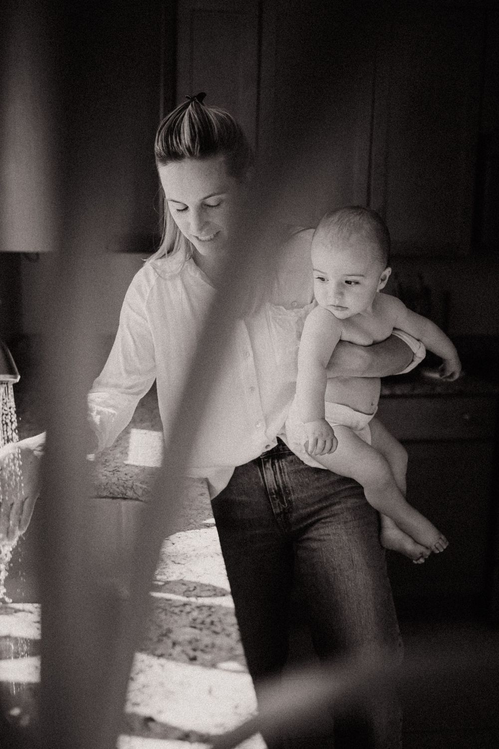 Black and white photo of a smiling mother holding her baby in a sunlit kitchen, wearing a white shirt and jeans.