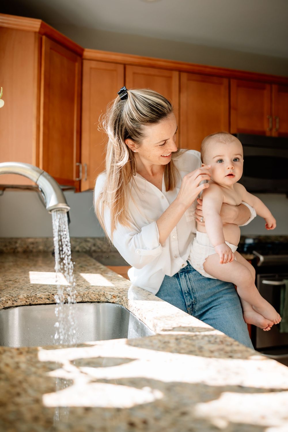 A young mother holds her baby near a kitchen sink with running water, smiling warmly in a sunlit home kitchen.