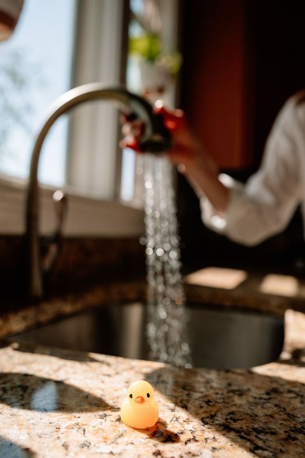 Rubber duck toy on granite countertop near kitchen sink with water flowing from faucet in sunlit kitchen.