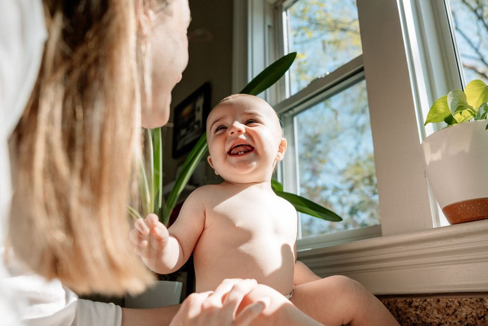 Happy baby laughing while being held near green tropical leaves by a window in bright natural light.