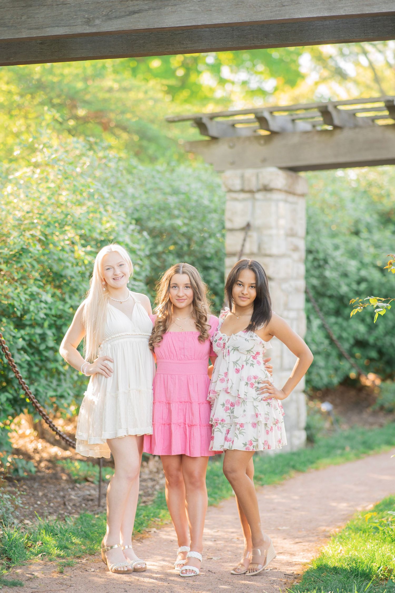 Three teenage girls in summer dresses posing under a garden pergola surrounded by greenery.