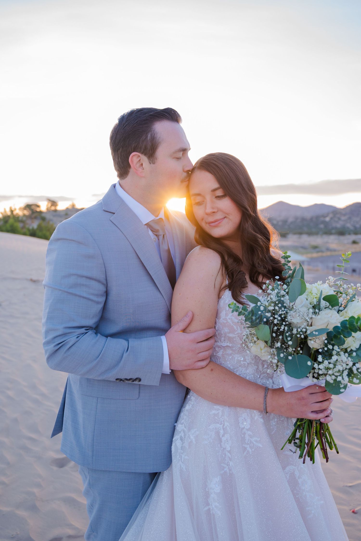 Groom in gray suit kisses bride on cheek at sunset on sandy desert dunes as she holds a white bouquet.
