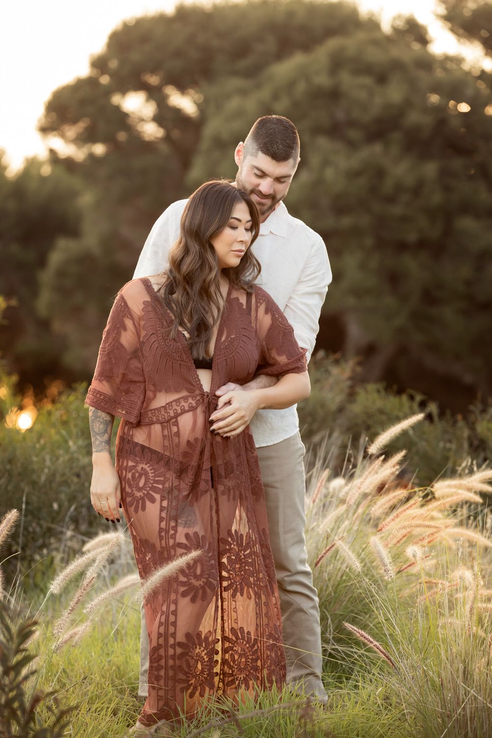 Couple embracing in golden hour light, woman in lace boho maternity dress, man in white shirt, surrounded by tall grass.