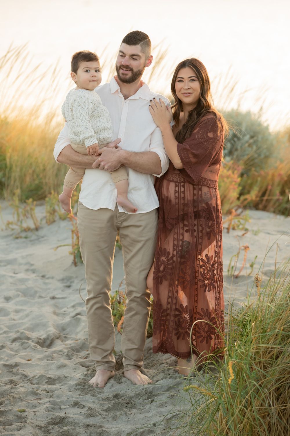 A young family poses on sandy beach dunes at golden hour, father holding baby, mother in rust floral dress.