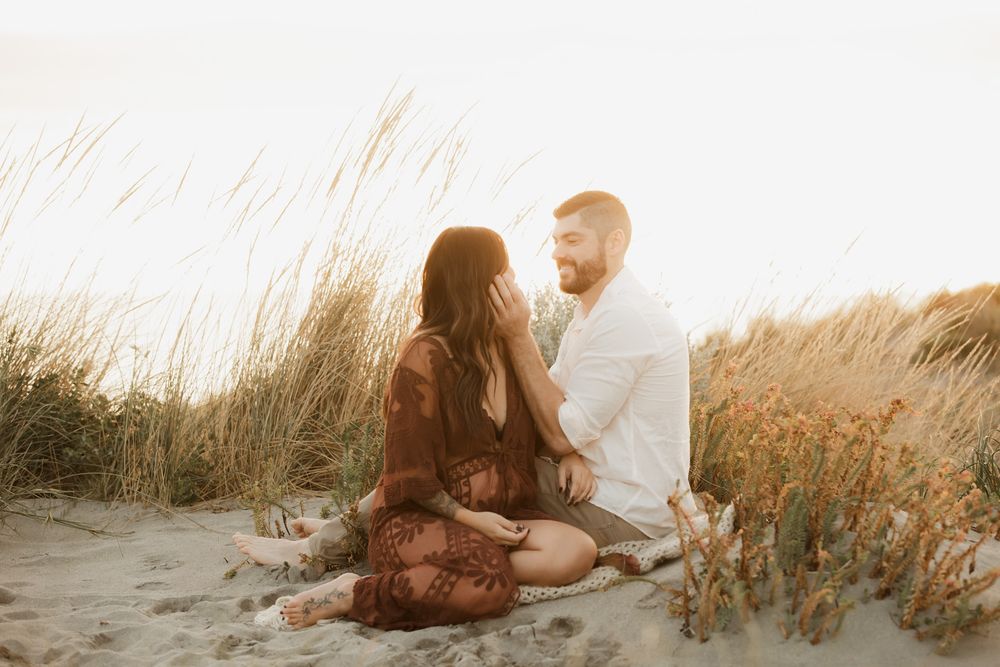 Couple sitting together on sandy beach dunes surrounded by golden grass in warm sunset light.