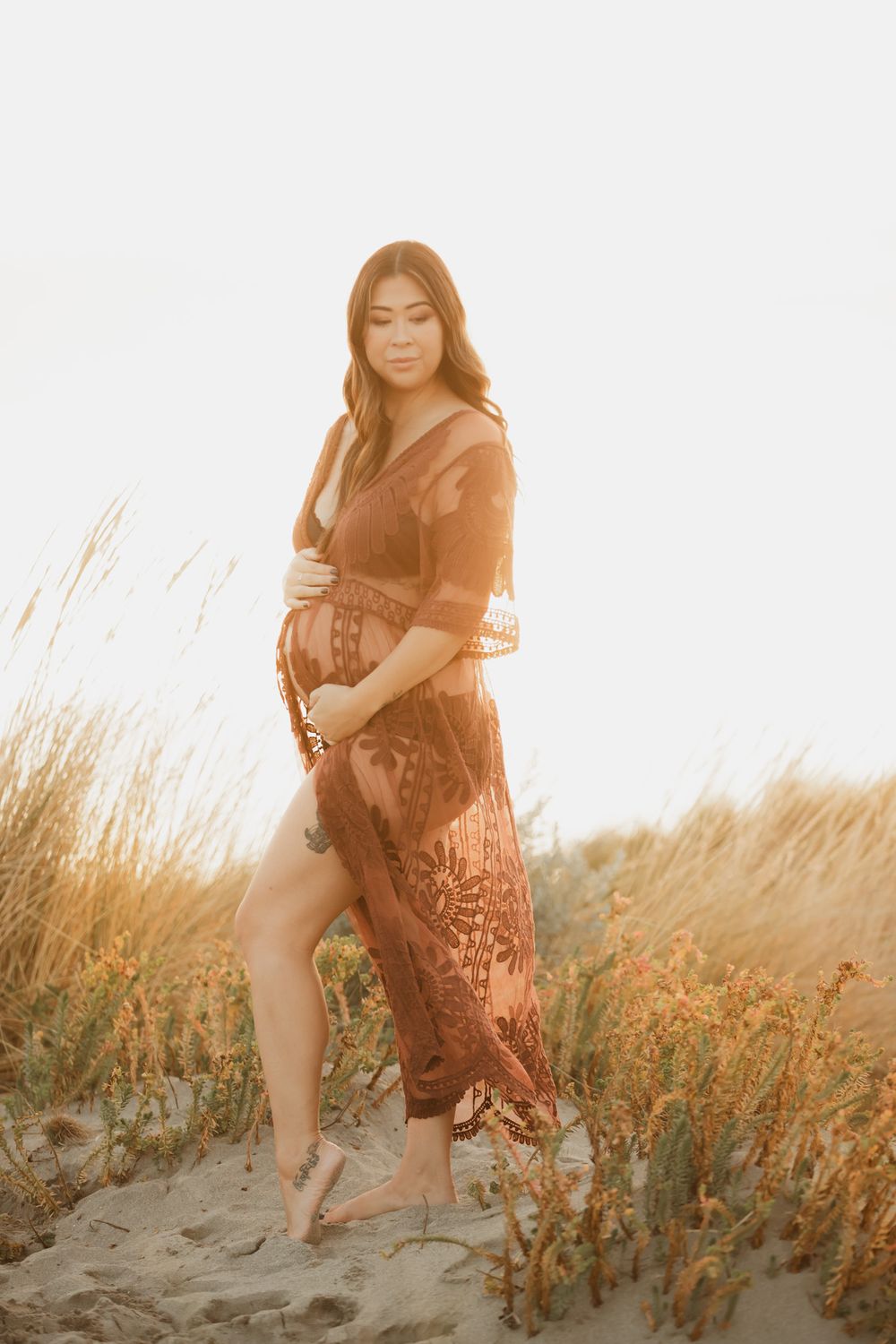 Pregnant woman in rust lace maxi dress standing barefoot on sandy dunes surrounded by golden grass at sunset.