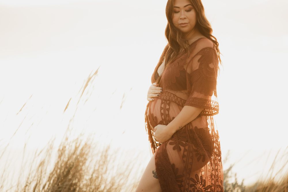 Pregnant woman in a rust lace dress cradling her belly outdoors in golden sunlight.