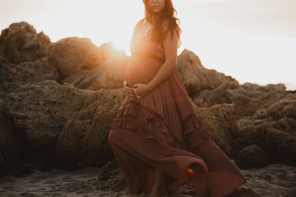 Pregnant woman in flowing rust gown poses on rocky beach at golden sunset.