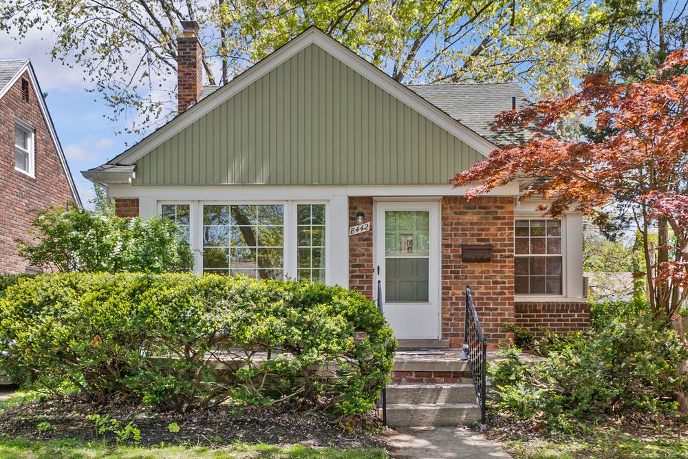 Charming brick bungalow with green siding, white front door, lush shrubs, and colorful Japanese maple tree.
