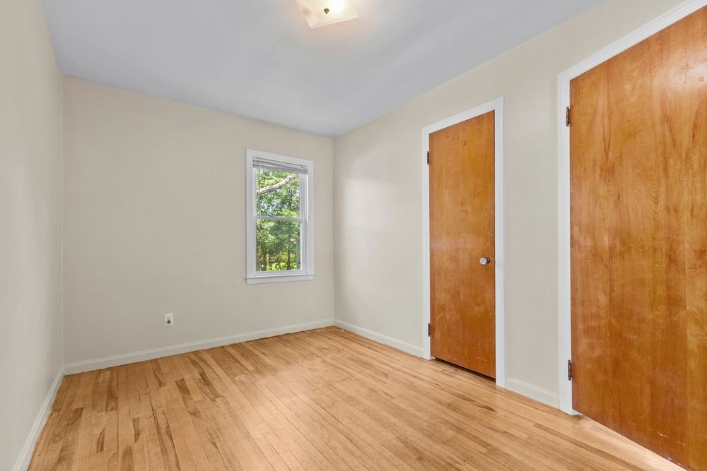Small empty bedroom with hardwood floors, single window with natural light, closet, and wood-tone door.