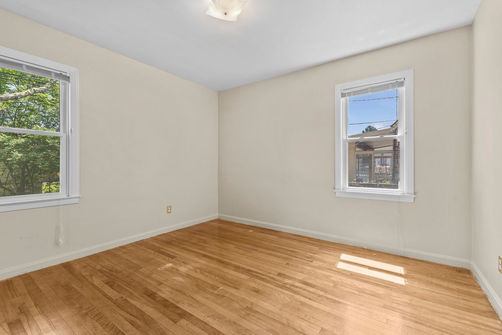 Bright empty bedroom with hardwood floors, two windows allowing ample natural light, and clean white walls.