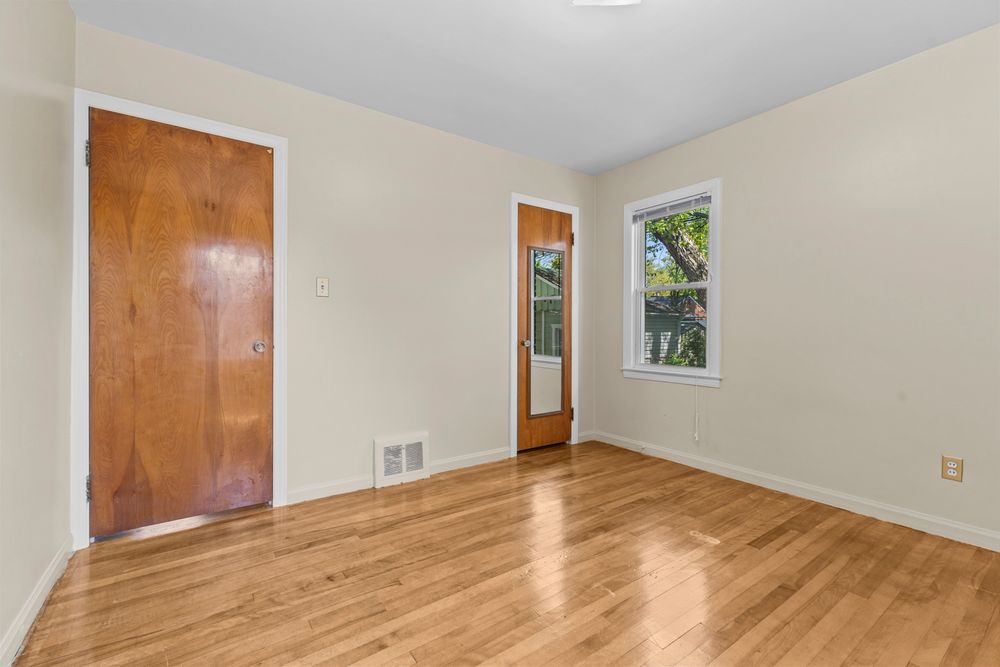 Empty bedroom with hardwood floors, two wood-tone doors, floor vent, and a window with natural light.