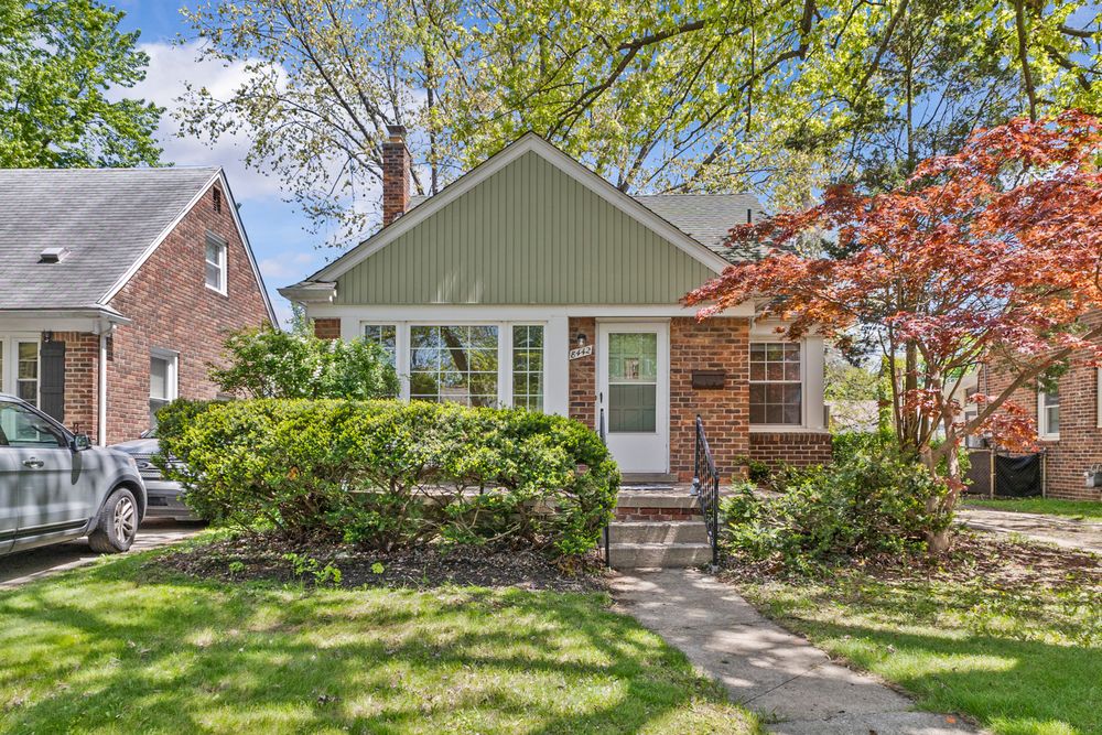 Charming brick bungalow with green siding, white front door, manicured shrubs, and vibrant Japanese maple.