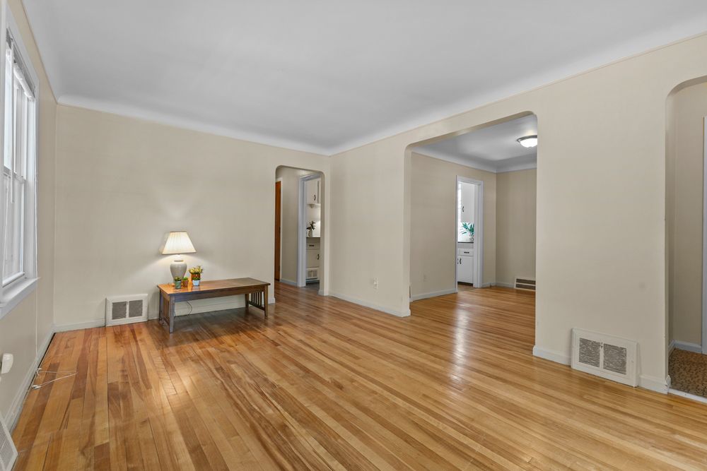 Bright empty living room with hardwood floors, crown molding, neutral walls, and open doorways to adjacent rooms.