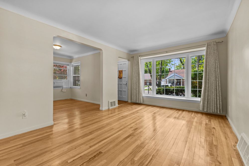 Spacious living room featuring gleaming hardwood floors, crown molding, large window with curtains, and open flow to adjacent room.