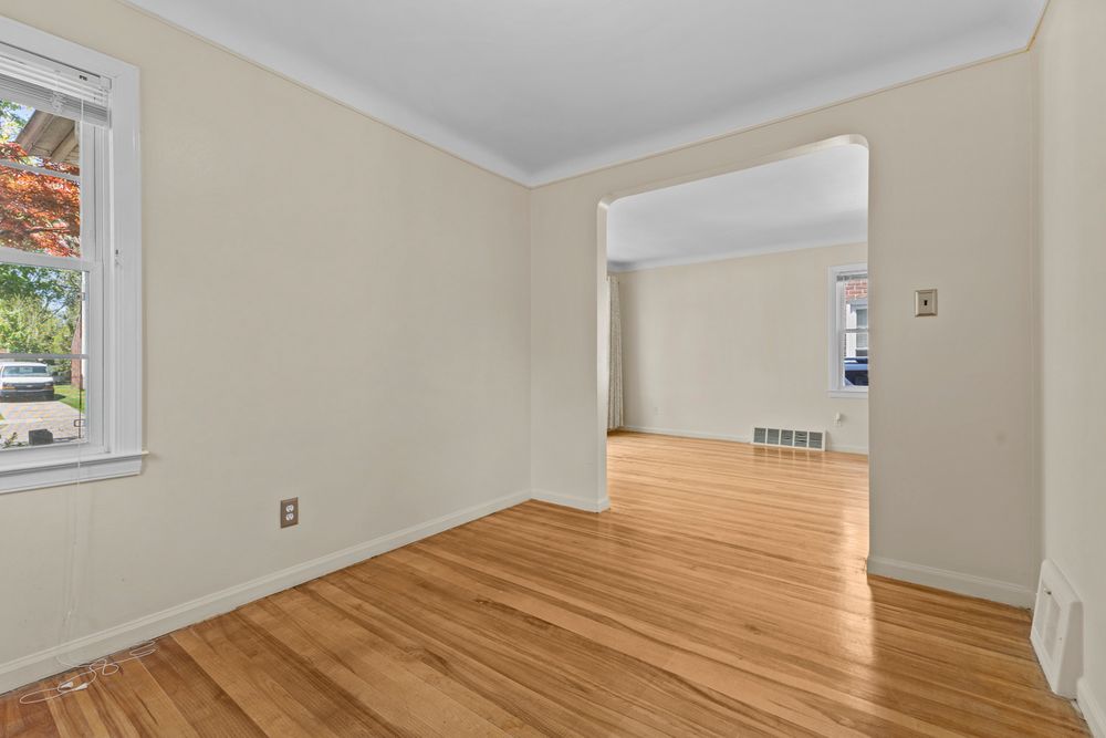 Vacant bedroom with hardwood floors, crown molding, natural light from window, and open doorway to bright living area.