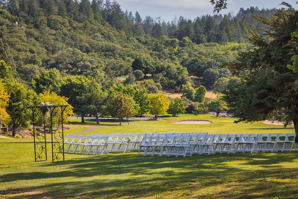 Outdoor wedding ceremony setup with white chairs on green lawn surrounded by trees and rolling hills.
