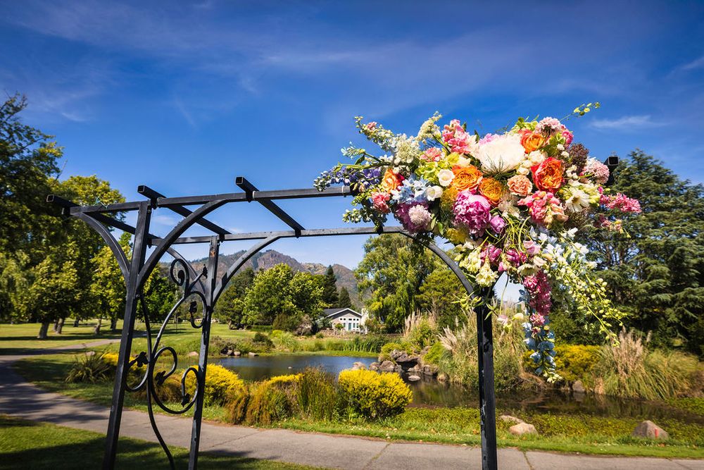 Colorful floral arrangement on black iron wedding arch in lush green park with pond and blue sky.