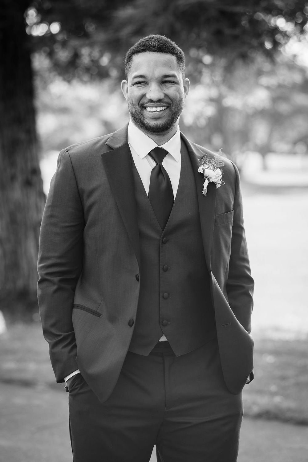 Smiling groom in a dark three-piece suit with boutonniere poses outdoors in black and white wedding photo.