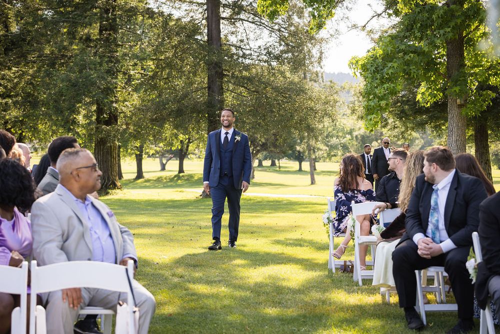 Groom stands at outdoor wedding ceremony aisle surrounded by seated guests on sunny day.