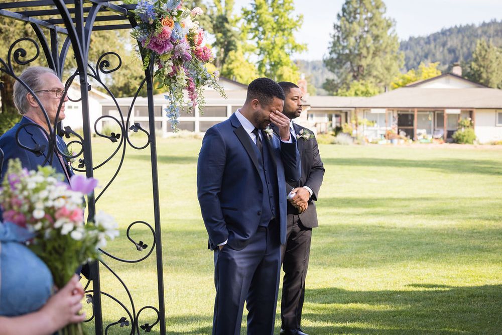 Two men in navy suits stand at an outdoor wedding ceremony altar decorated with flowers and iron arch.