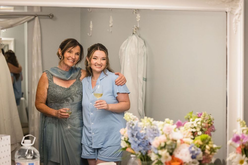 Two women smiling in a mirror reflection, one in a blue bridesmaid dress holding champagne at a wedding.