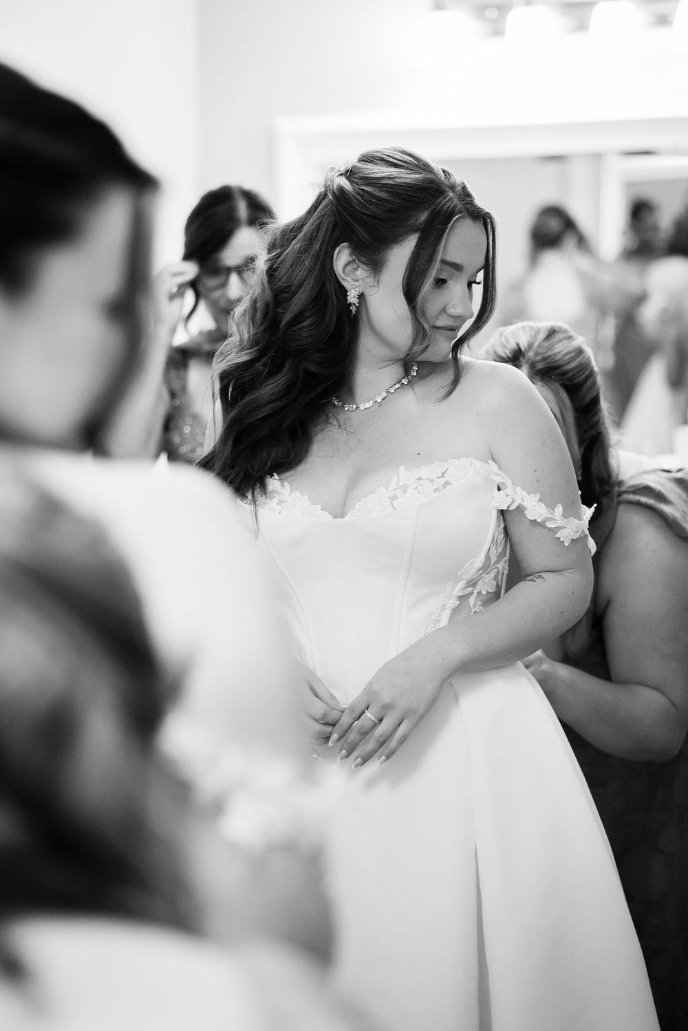 Black and white photo of a bride in an off-shoulder wedding gown being helped by attendants in a bridal suite.