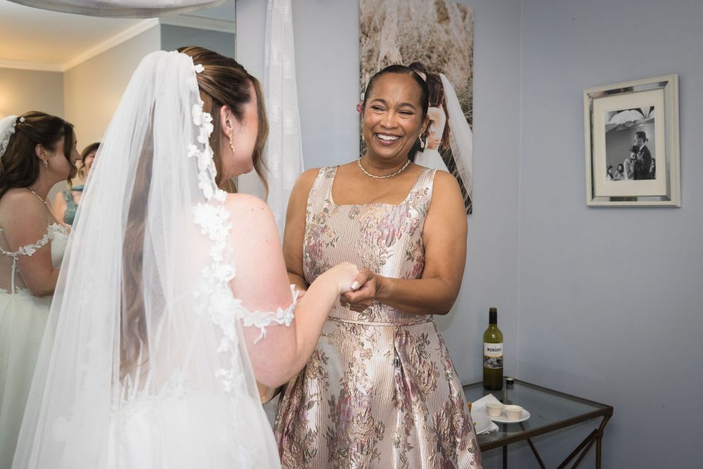 Bride in white lace gown and veil shares a joyful moment with smiling bridesmaid in elegant gold dress.