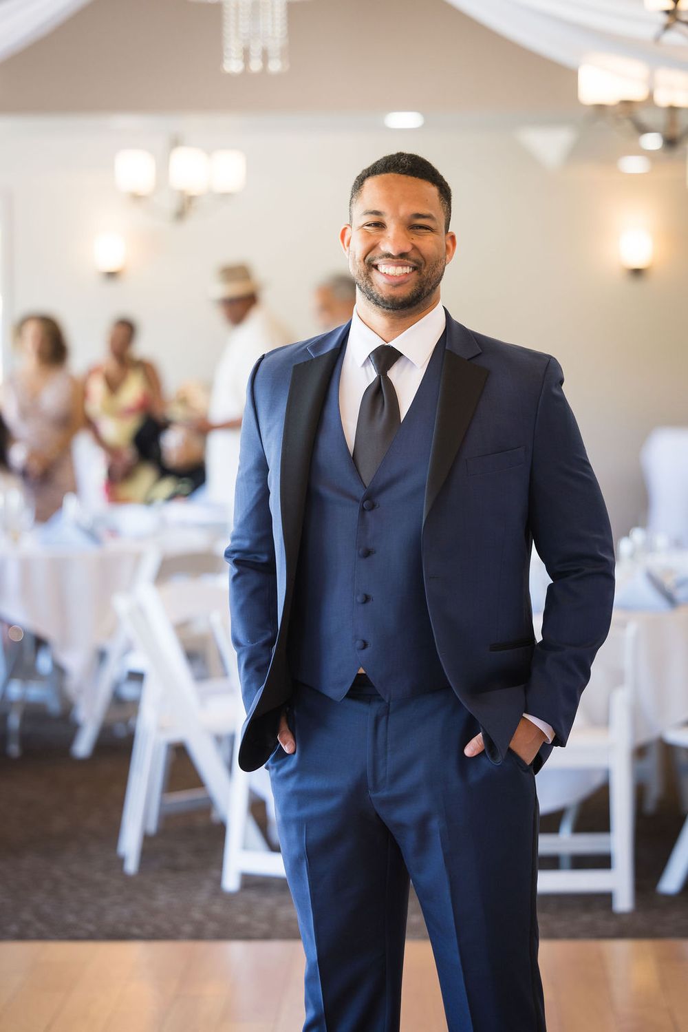 Handsome groom in a navy blue three-piece suit smiling at wedding reception venue.