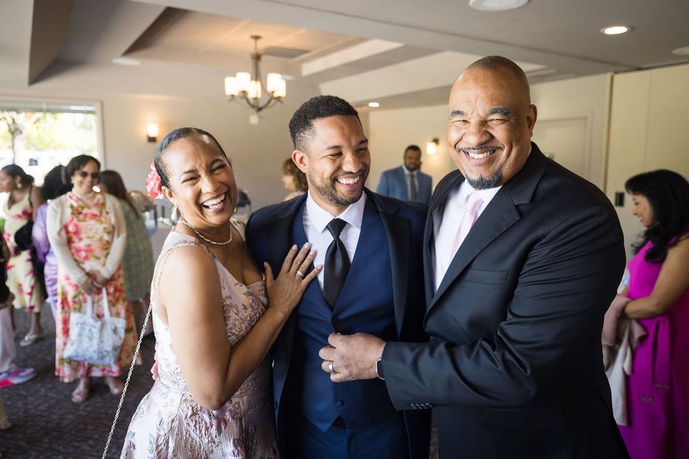 Groom in navy suit laughs with family members during joyful wedding reception celebration.