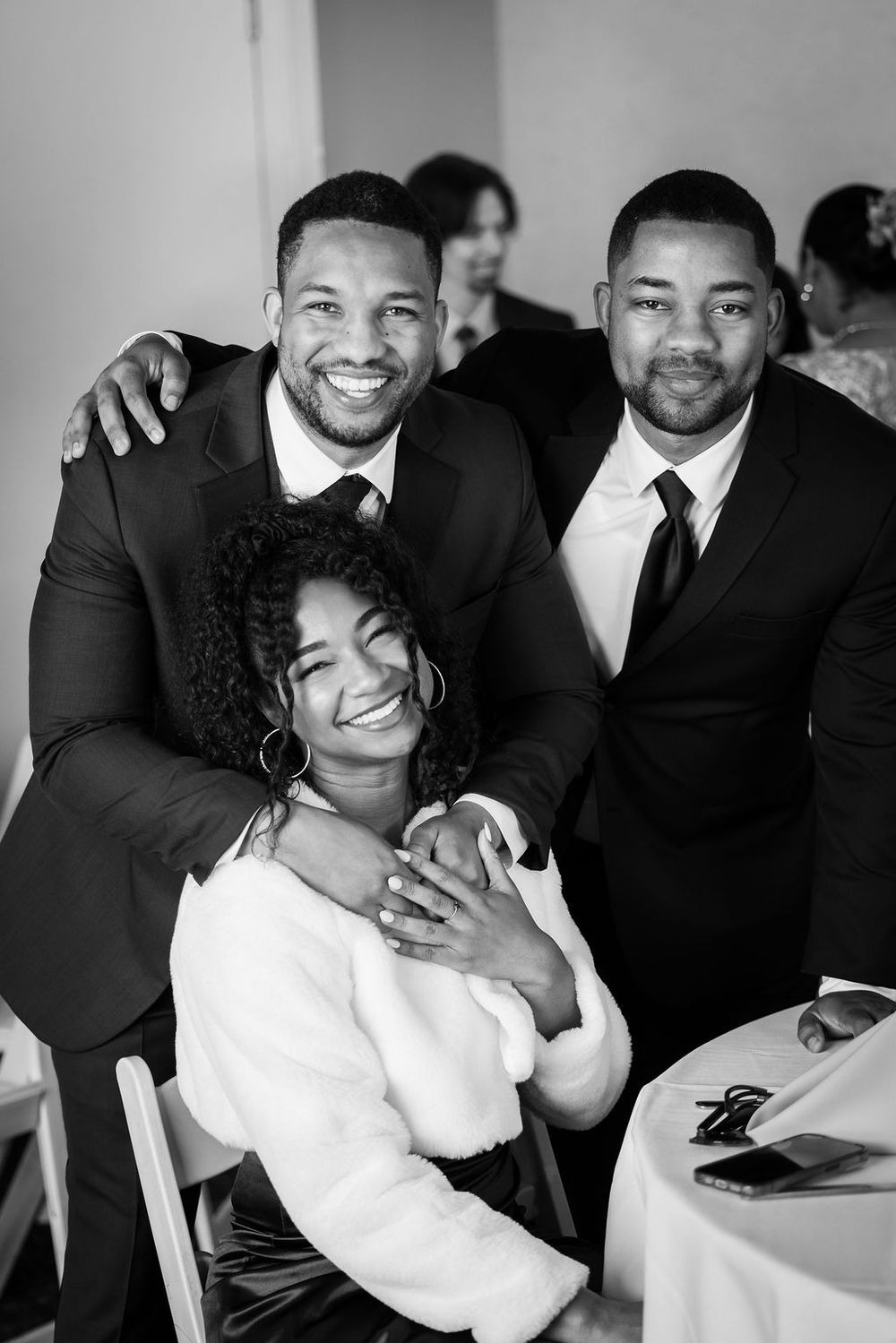 Two men in suits smile with a woman in a white fur wrap at a wedding reception in black and white.