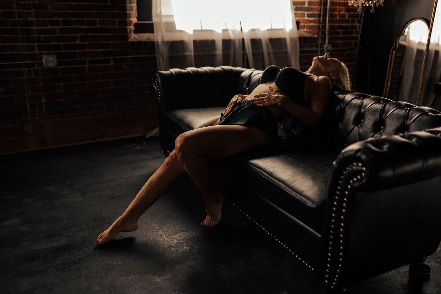 Person wearing black strappy lingerie posing in dramatic low-key lighting on a dark surface.