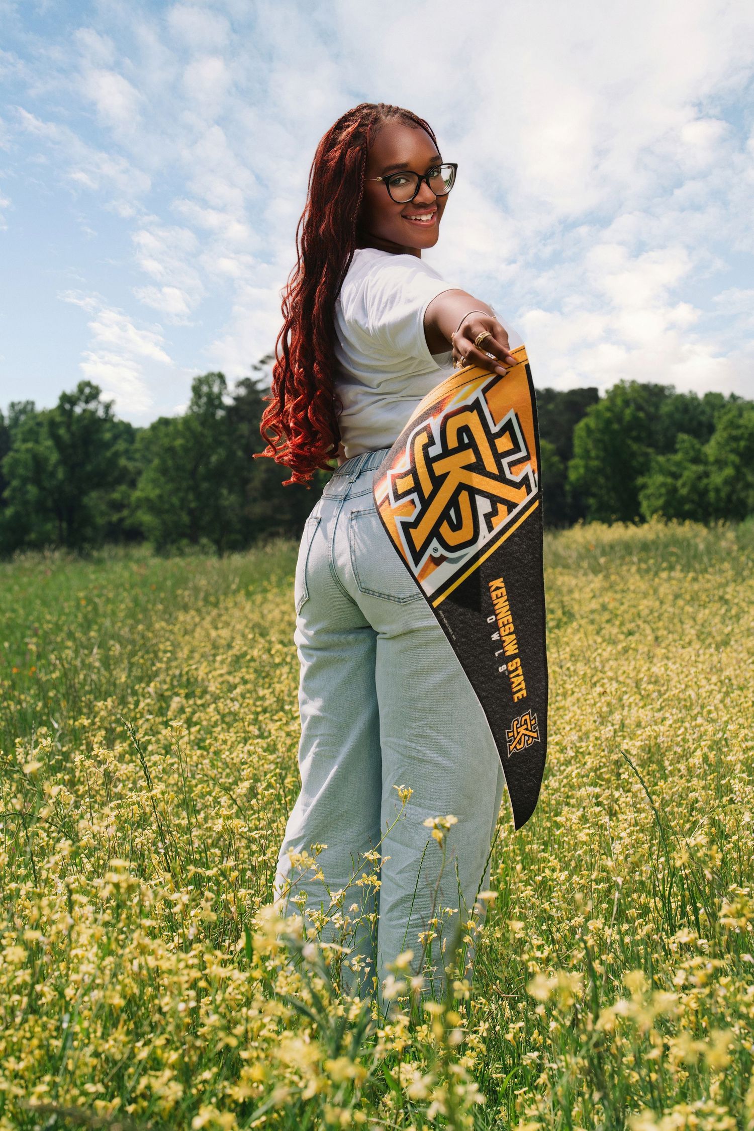 Young woman holding a black and gold college pennant flag standing in a sunny wildflower field, smiling over her shoulder.