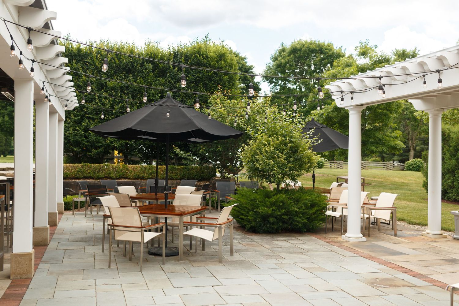 A charming white farmhouse surrounded by lush green trees and outdoor seating on a sunny day.