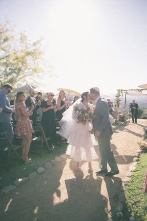 Bride and groom share a kiss while walking down the aisle at an outdoor wedding ceremony on a sunny day.