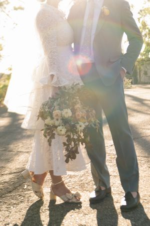 Bride and groom standing together outdoors in golden sunlight, bride holding floral bouquet in lace dress and heels.