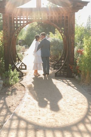 Bride and groom kissing under a circular wooden garden arch, with sun flare and shadows on a sandy path.