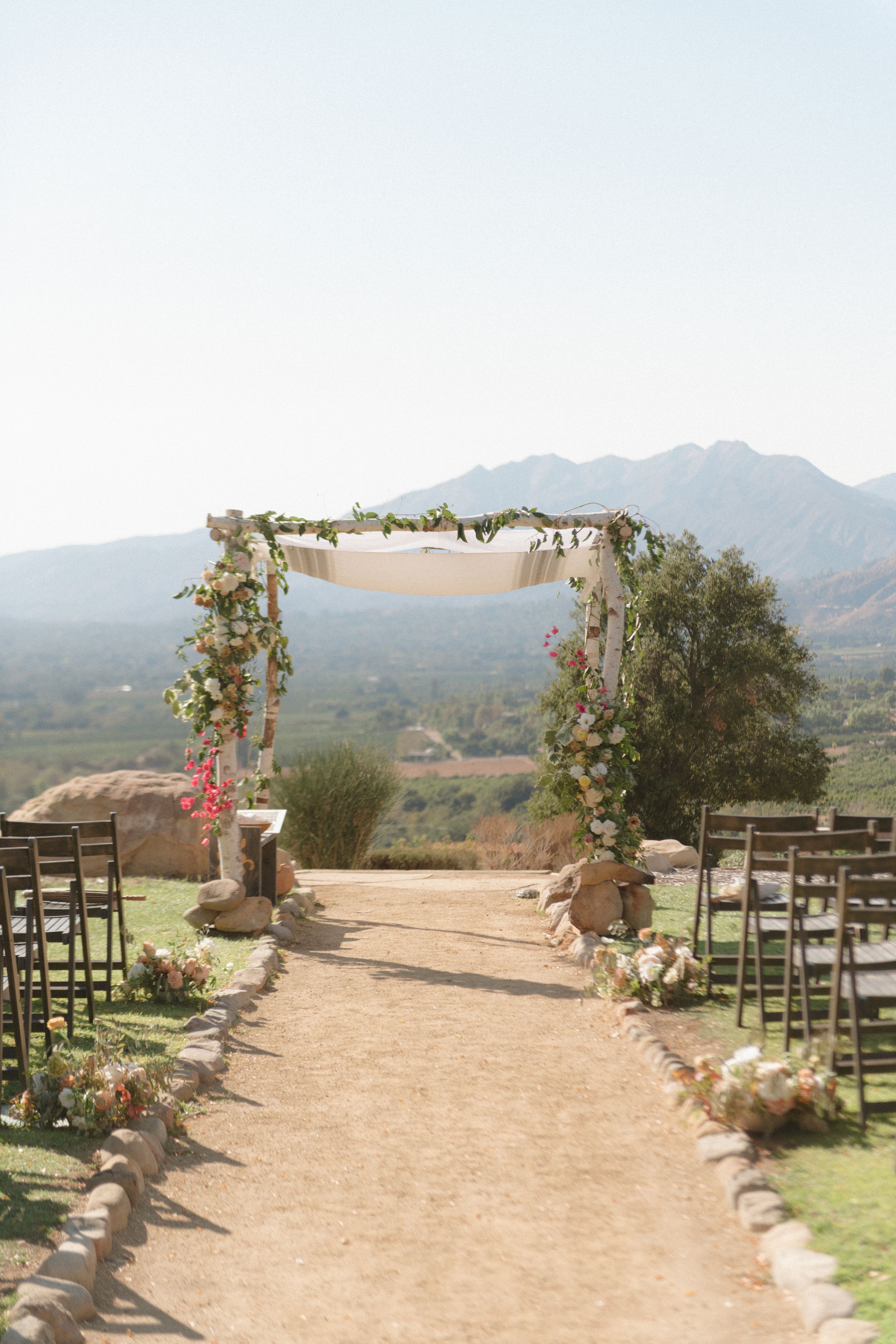 Outdoor wedding ceremony aisle with floral arch, wooden chairs, and stunning mountain backdrop under clear blue sky.