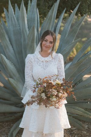 Bride in lace long-sleeve wedding dress holding autumn bouquet, posing in front of large agave plant outdoors.